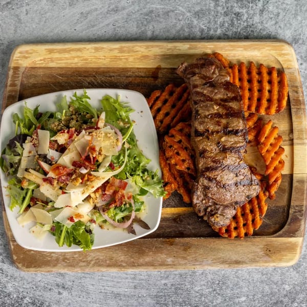 Grilled steak with waffle fries and a fresh salad topped with cheese and bacon on a wooden board.