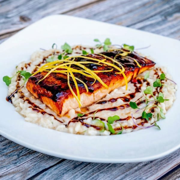 Seared salmon with balsamic glaze and lemon zest on a bed of creamy risotto, garnished with microgreens.
