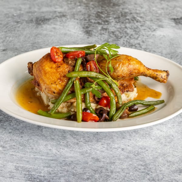 Golden-brown roasted chicken leg and thigh on mashed potatoes with green beans, tomatoes, and olives.