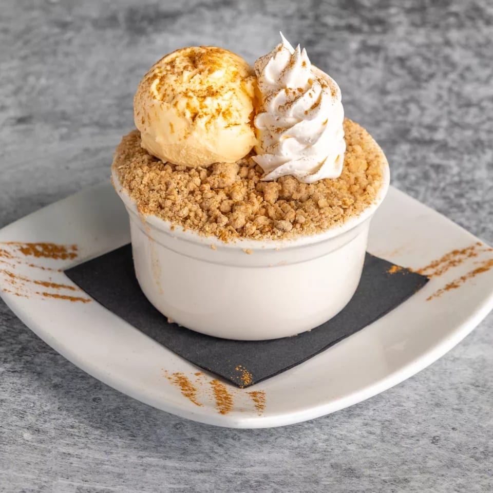 A dessert in a white bowl with crumble, a scoop of ice cream, and whipped cream.