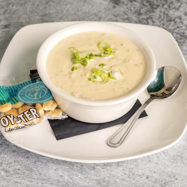 Creamy clam chowder in a white bowl, garnished with green onions, served with oyster crackers and a spoon.
