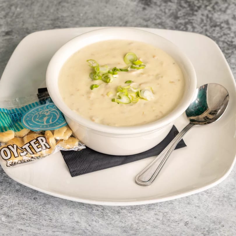 Creamy clam chowder in a white bowl, garnished with green onions, served with oyster crackers and a spoon.