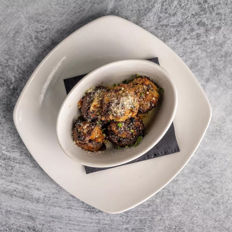 Top-down view of a white bowl filled with five stuffed mushrooms, sprinkled with cheese and herbs.