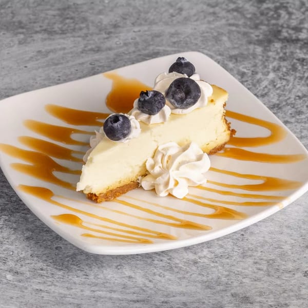 Slice of cheesecake with blueberries, whipped cream, and caramel drizzle on a white plate.