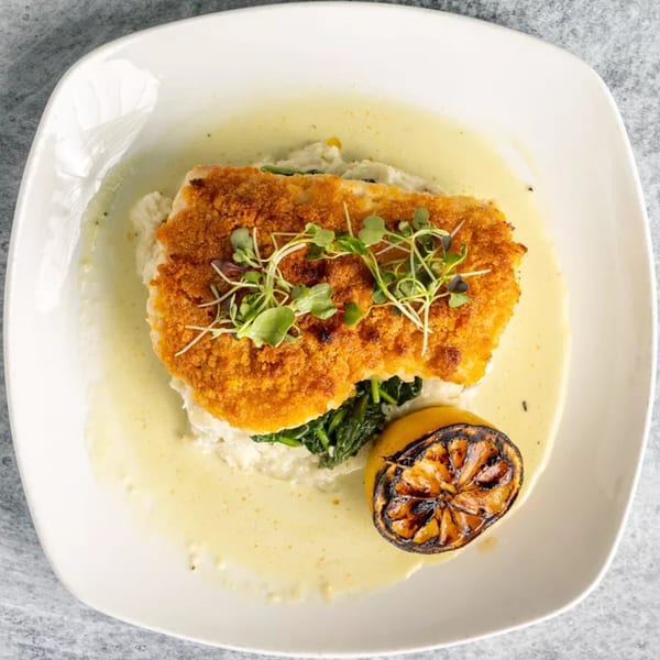 Crispy breaded fish with microgreens, spinach, mashed potatoes, sauce, and a grilled lemon.
