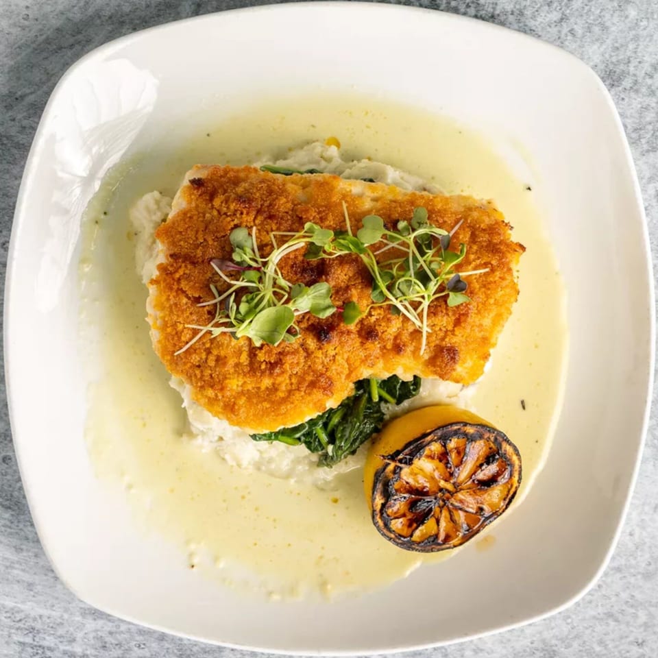 Crispy breaded fish with microgreens, spinach, mashed potatoes, sauce, and a grilled lemon.