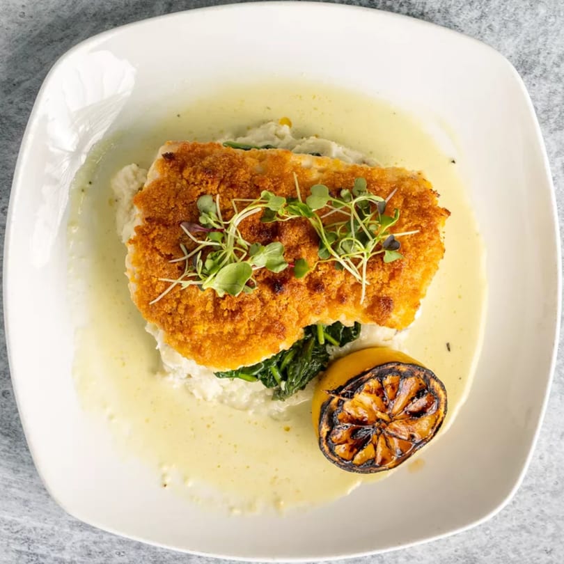 Crispy breaded fish with microgreens, spinach, mashed potatoes, sauce, and a grilled lemon.