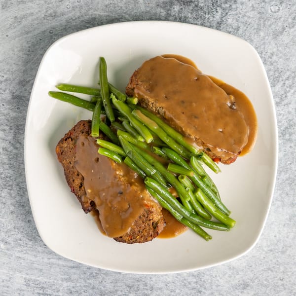 Two slices of meatloaf covered in brown gravy, served with a side of green beans on a white plate.