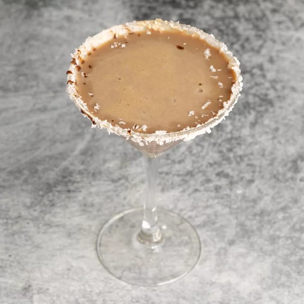A creamy brown cocktail in a martini glass with a coconut-flaked rim, against a gray background.