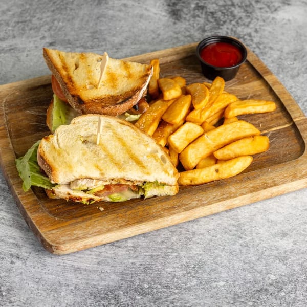 Grilled chicken sandwich with lettuce, tomato, and avocado, served with steak fries and ketchup on a wooden board.